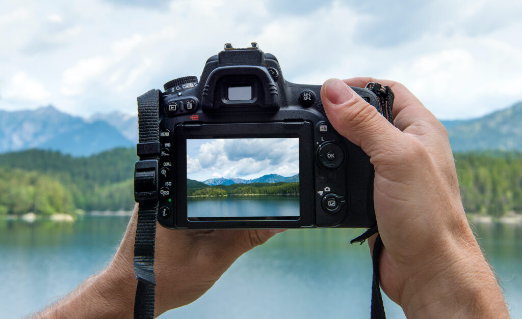 Hands of a male photographer holding a digital camera taking pictures of a idyllic landscape with a lake and mountains while the picture shows at the display